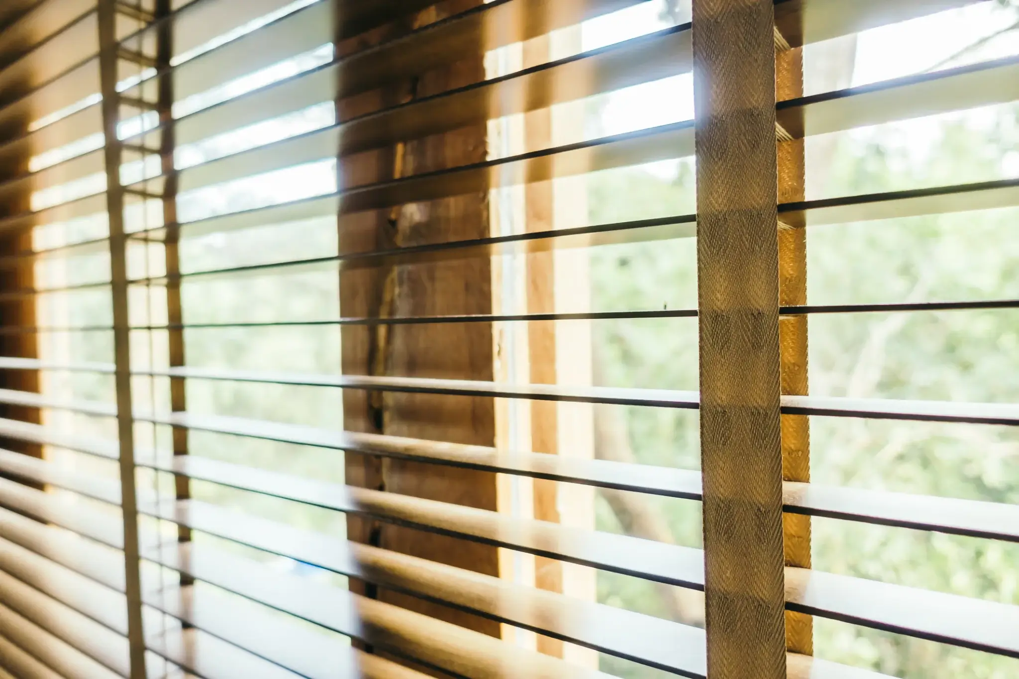 Drewniane żaluzje okienne na tle zieleni – elegancka i naturalna osłona wnętrza chroniąca przed słońcem.